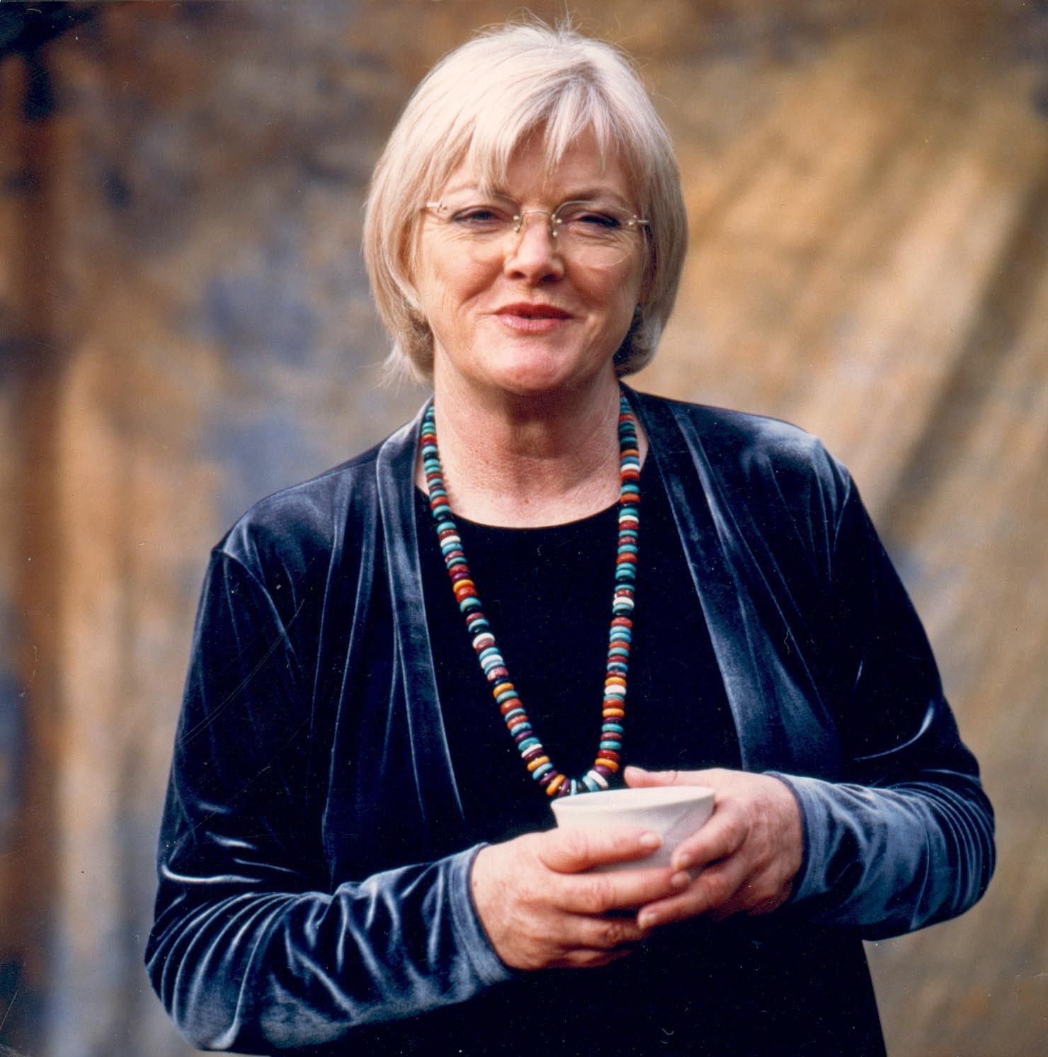 Portrait of a smiling person in a blue velvet jacket, holding a white cup against a soft studio backdrop