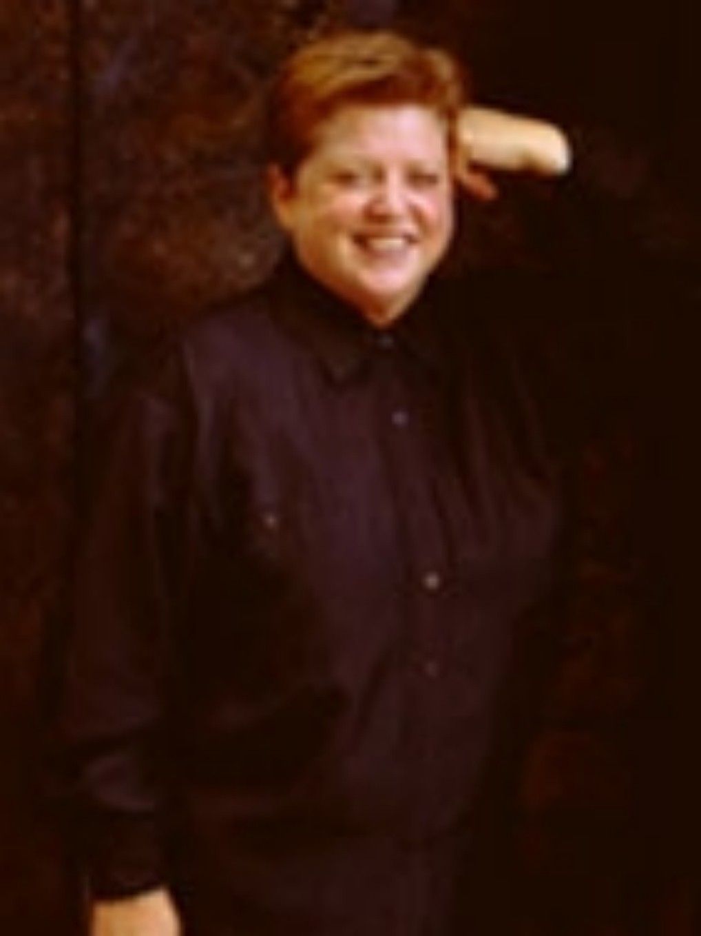 Smiling person in a dark button-up shirt standing against a brown background