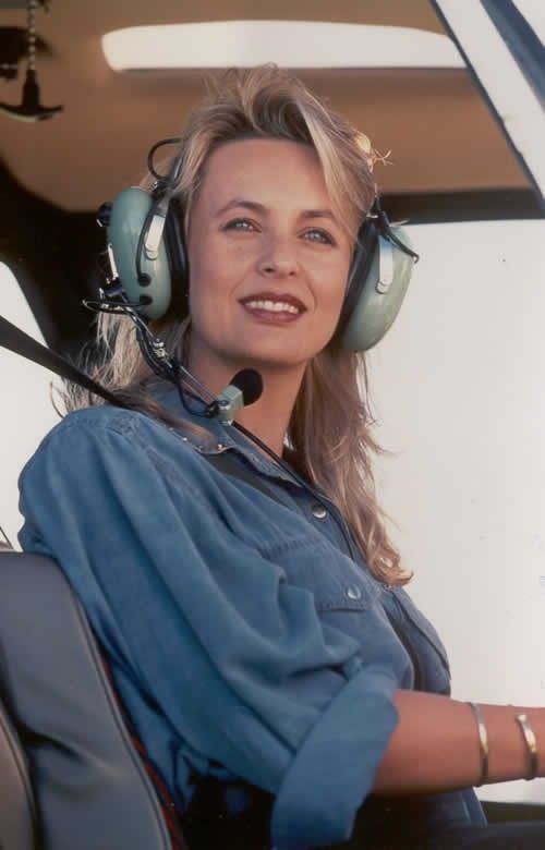 Woman in a helicopter cockpit wearing a headset and blue shirt, smiling slightly.