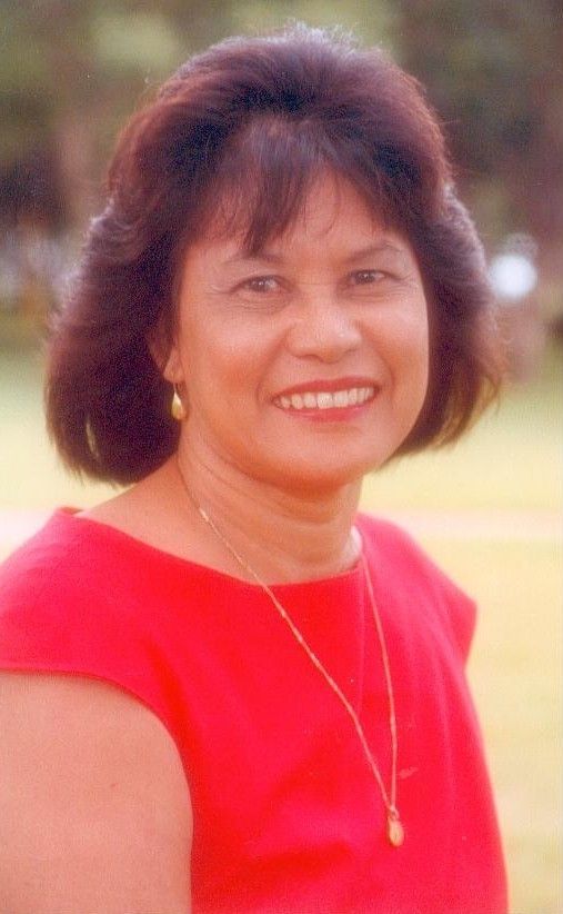 Smiling woman in a bright red top with short dark hair, standing outdoors against a blurred background