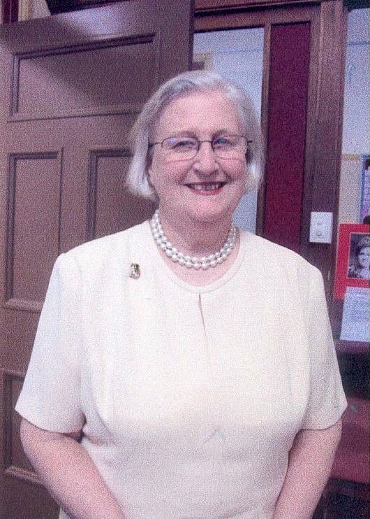 Smiling woman in a light cream top and pearl necklace standing indoors by a wooden door