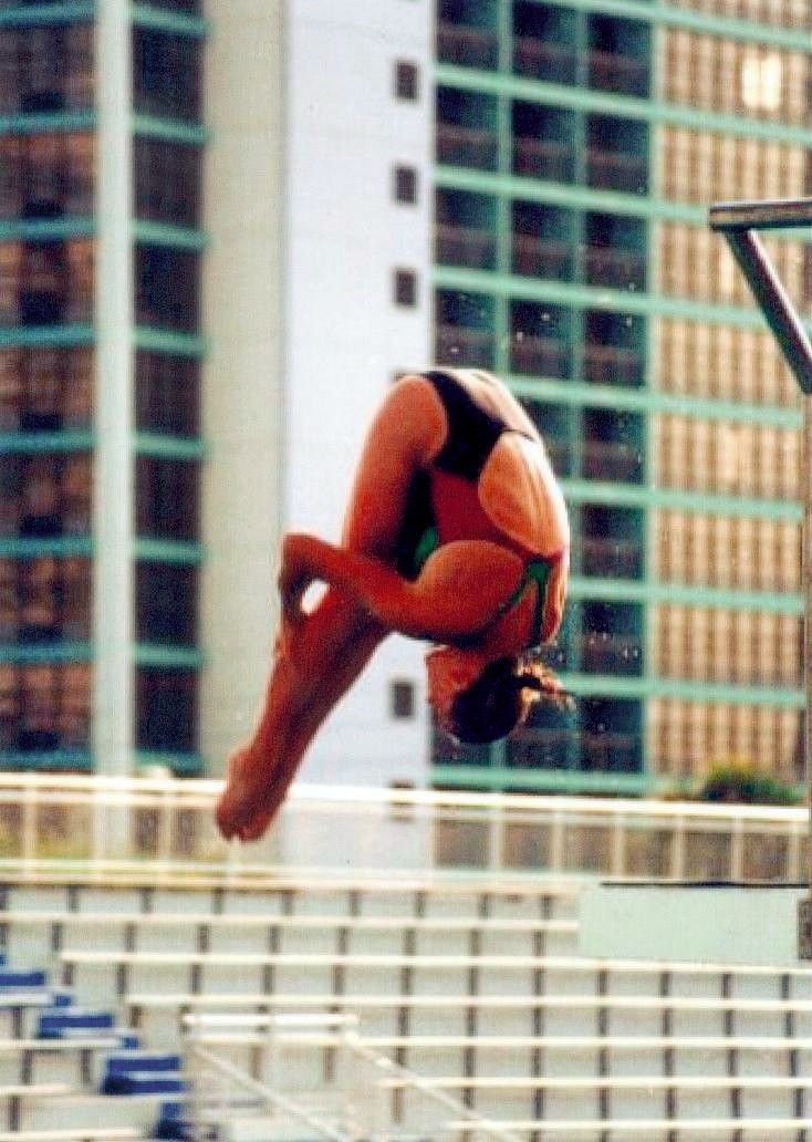 Gymnast in orange leotard performing a midair flip near bleachers and a tall building