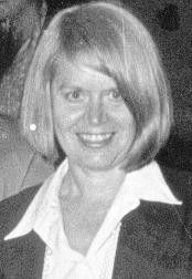 Smiling woman with short blonde hair in a dark jacket and white collared shirt.