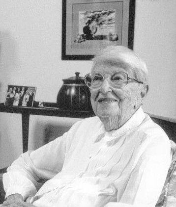 Smiling elderly woman in glasses and white blouse sitting indoors by a table with framed art behind her