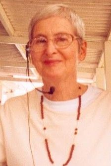 Smiling person with short gray hair and glasses, wearing a white shirt and necklace indoors.