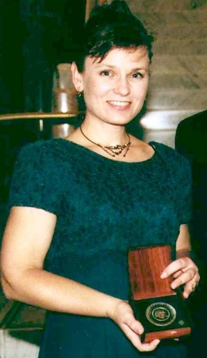 Woman in a dark green dress holding a small award plaque and smiling indoors