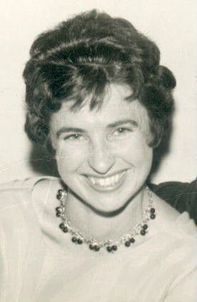 Smiling person with short curly dark hair and a necklace, posing against a plain background.