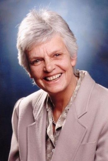 Smiling person with short white hair in a light blazer against a blue studio backdrop