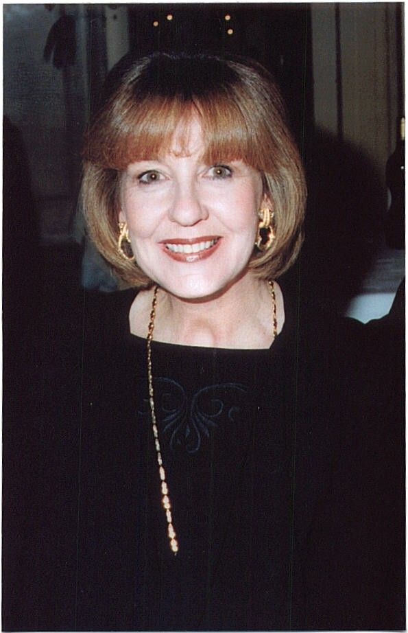 Smiling woman with short blonde hair wearing a black top and gold jewelry indoors