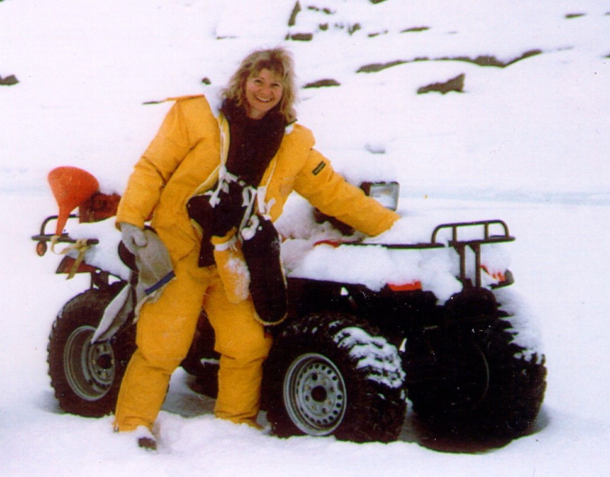 Person in yellow snowsuit smiling beside a snow-covered ATV in a snowy field