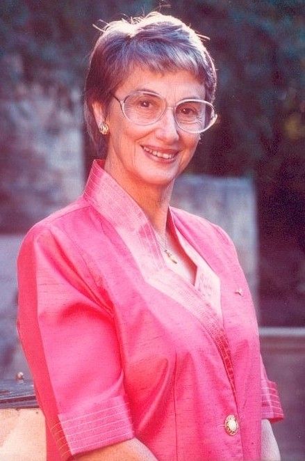 Smiling woman in a pink blazer and glasses standing outdoors against a blurred background