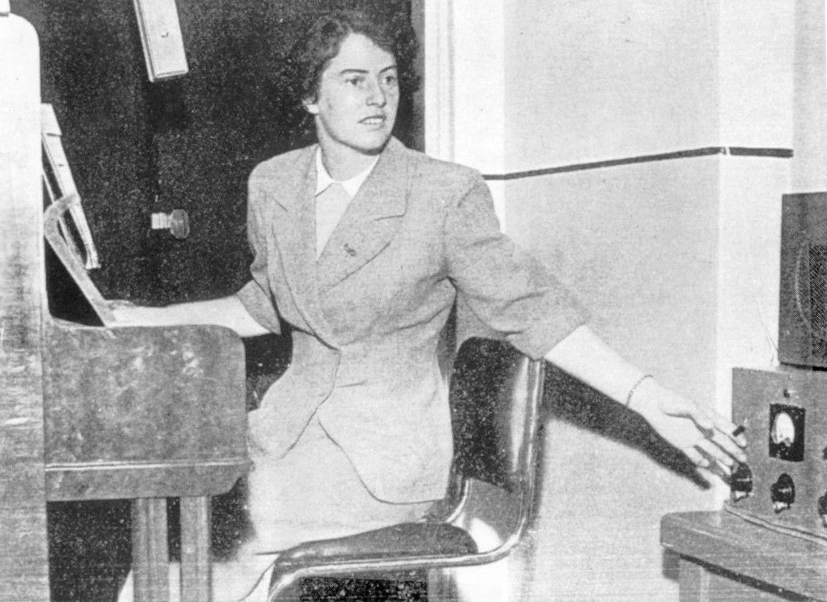 Woman seated at a piano, turning toward a control panel in a black-and-white photo