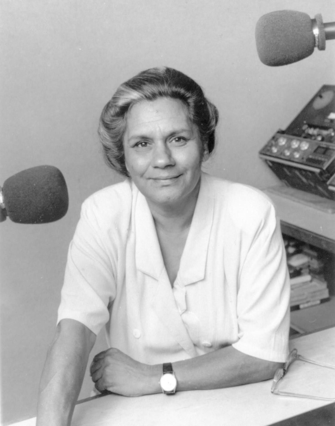 Woman in a radio studio, seated at a desk with microphones and audio equipment, smiling at the camera
