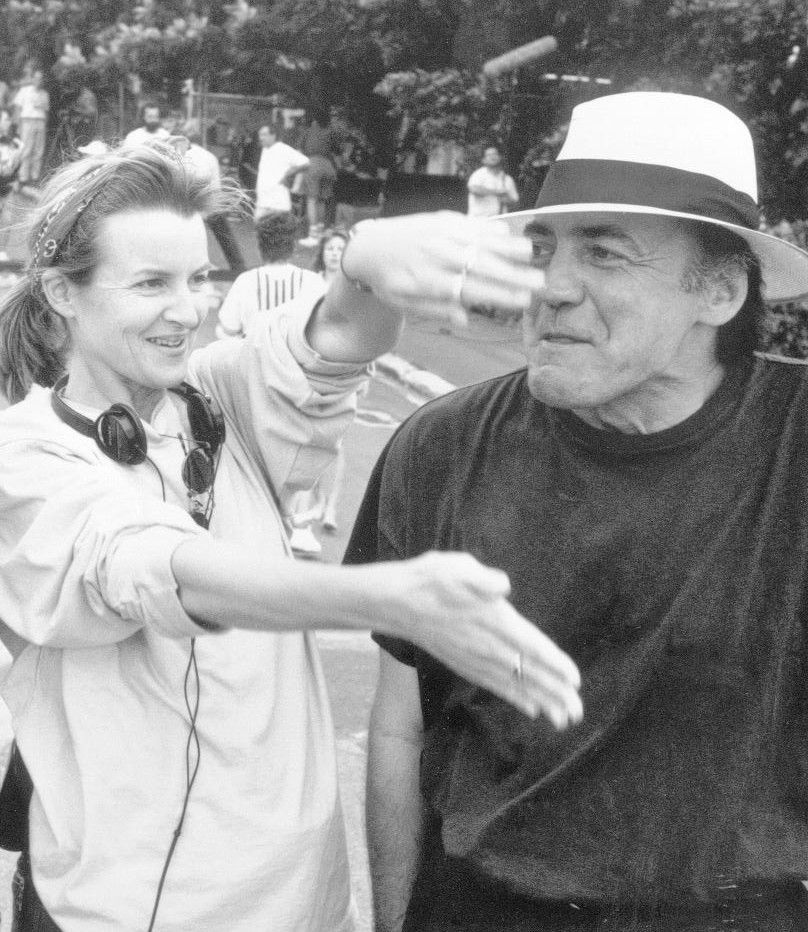 Two people smiling and gesturing outdoors, one wearing a white hat and the other with headphones around her neck.