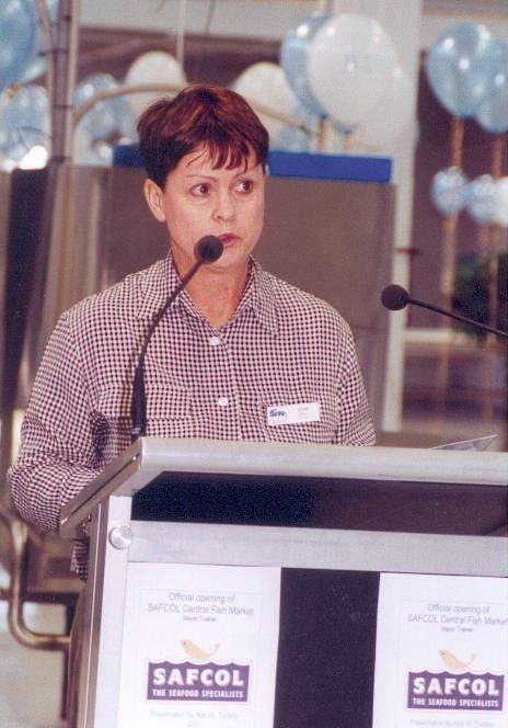 Woman speaking at a podium with microphones, in front of SAFCOL signage and balloons.