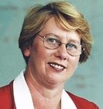 Portrait of a smiling person with short blond hair, glasses, and a red-and-white jacket against a blue background