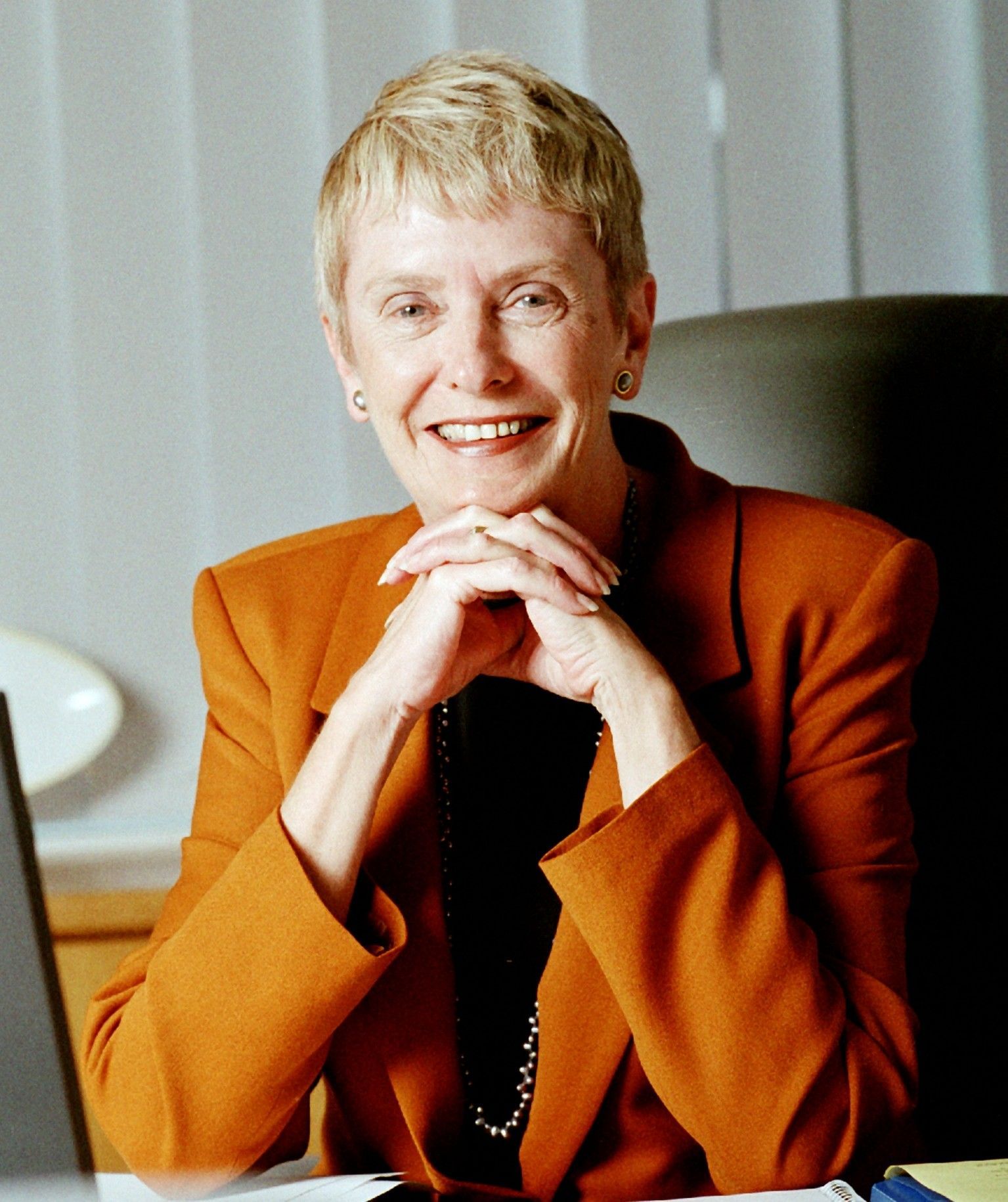 Smiling person in an orange blazer, seated with hands clasped against a light background.