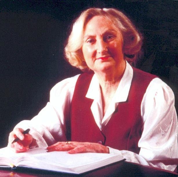 Woman in a white blouse and red vest writes in a notebook at a table against a dark background