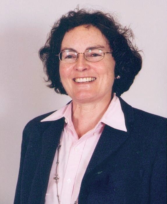 Portrait of a smiling person in glasses, wearing a dark blazer and pink shirt against a light background