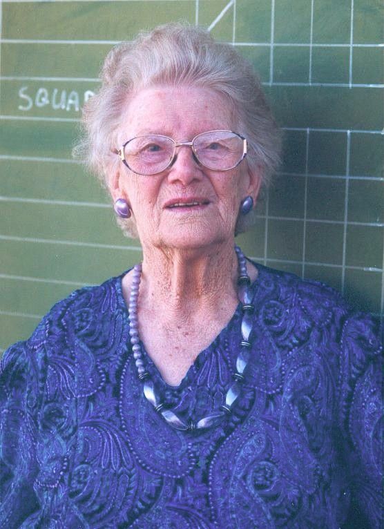 Elderly woman in glasses wearing a purple patterned blouse, standing in front of a chalkboard.