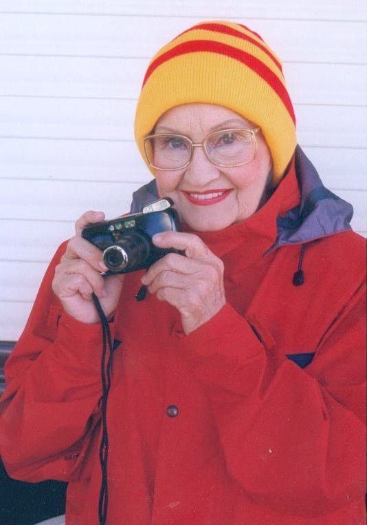 Smiling person in red coat and yellow hat holding a camera outdoors in snow
