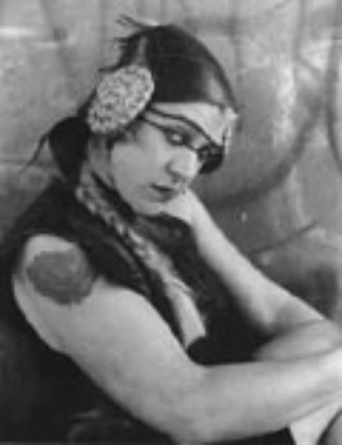 Black-and-white portrait of a person in theatrical costume with braided hair, headpiece, and crossed arms.