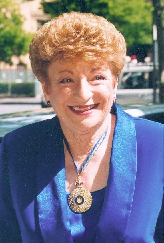 Smiling woman in a blue jacket and large pendant necklace outdoors in sunlight