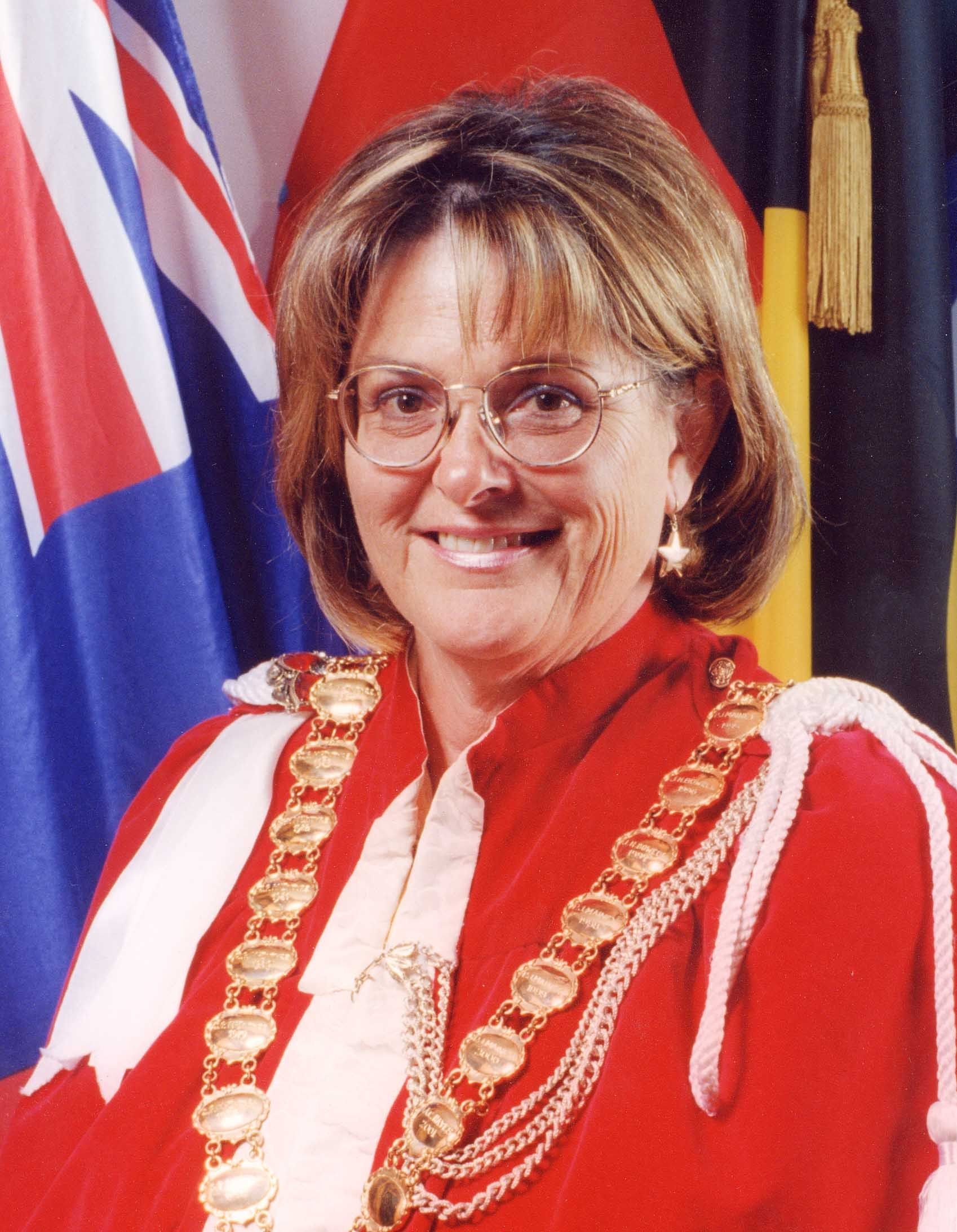 Woman in red ceremonial robe with gold chain, smiling in front of flags.
