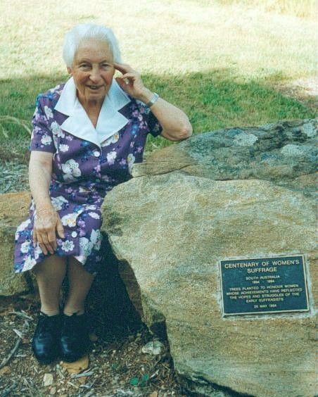 Elderly woman in a purple dress sitting on a rock beside a memorial plaque outdoors