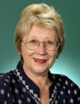 Portrait of a smiling woman with short blonde hair, glasses, and a dark patterned blouse on a green background