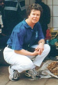 Person crouching outdoors in a blue shirt and white pants, holding a camera, with people in the background