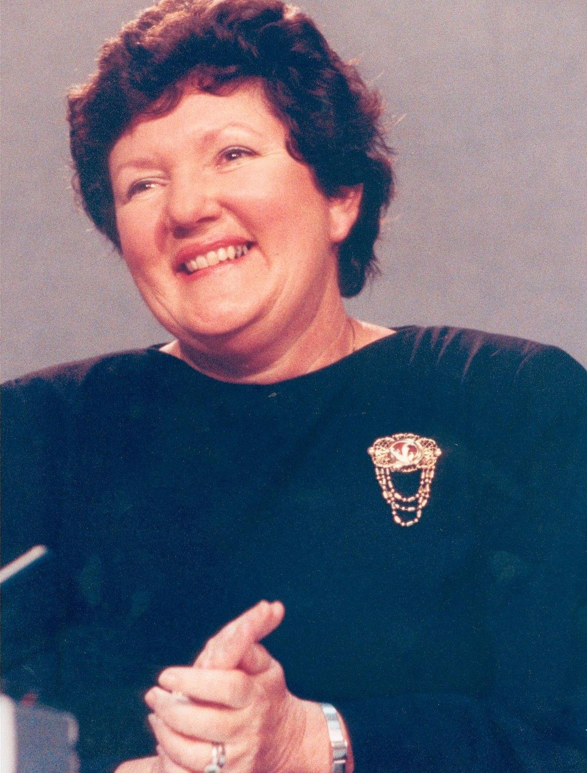 Smiling woman in a dark top with a brooch, hands clasped against a plain background