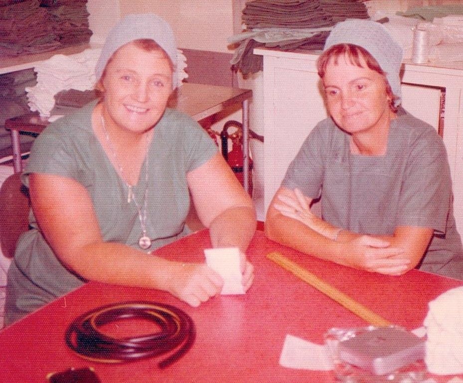 Two people in gray shirts and white bonnets sit at a red table with a cup and black coiled cord.