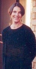 Smiling person in a black top standing indoors beside a window.