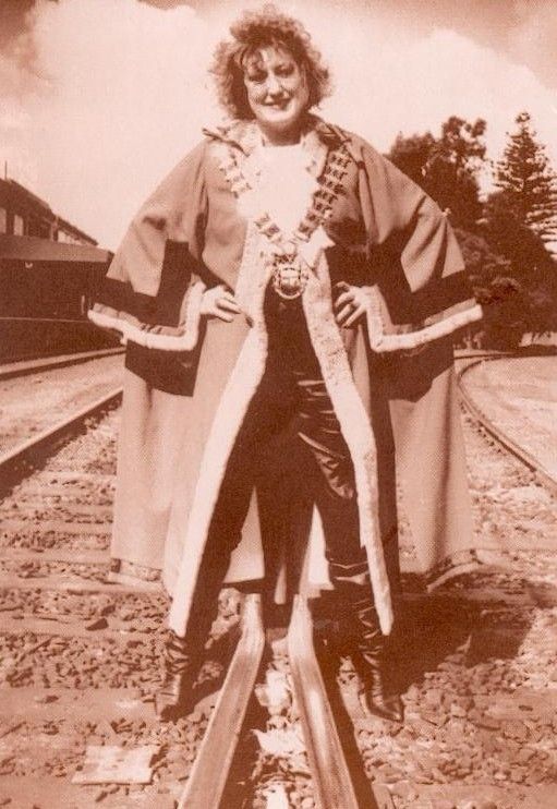 Person standing on railroad tracks in a flowing robe, smiling at the camera.