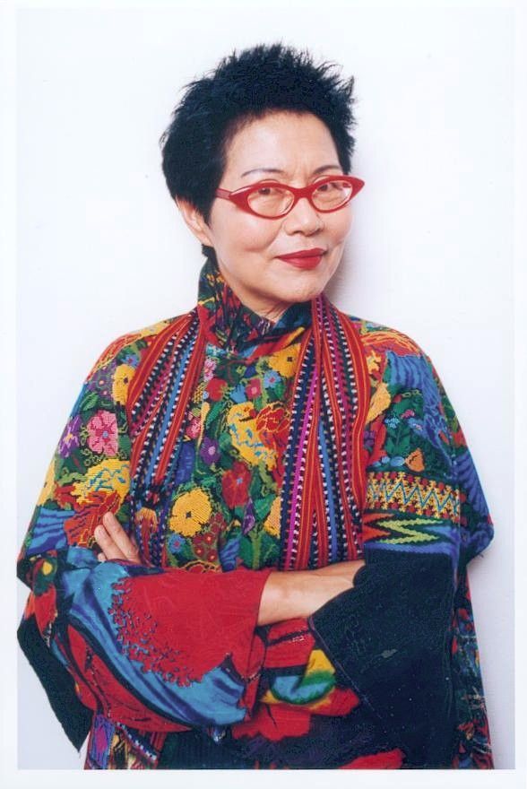 Portrait of a person in red glasses and a colorful patterned blouse, arms crossed against a white background