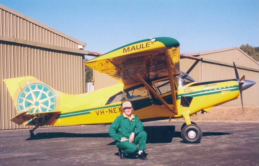 Yellow-green Maule airplane parked on pavement with a person kneeling in front of it by a hangar.