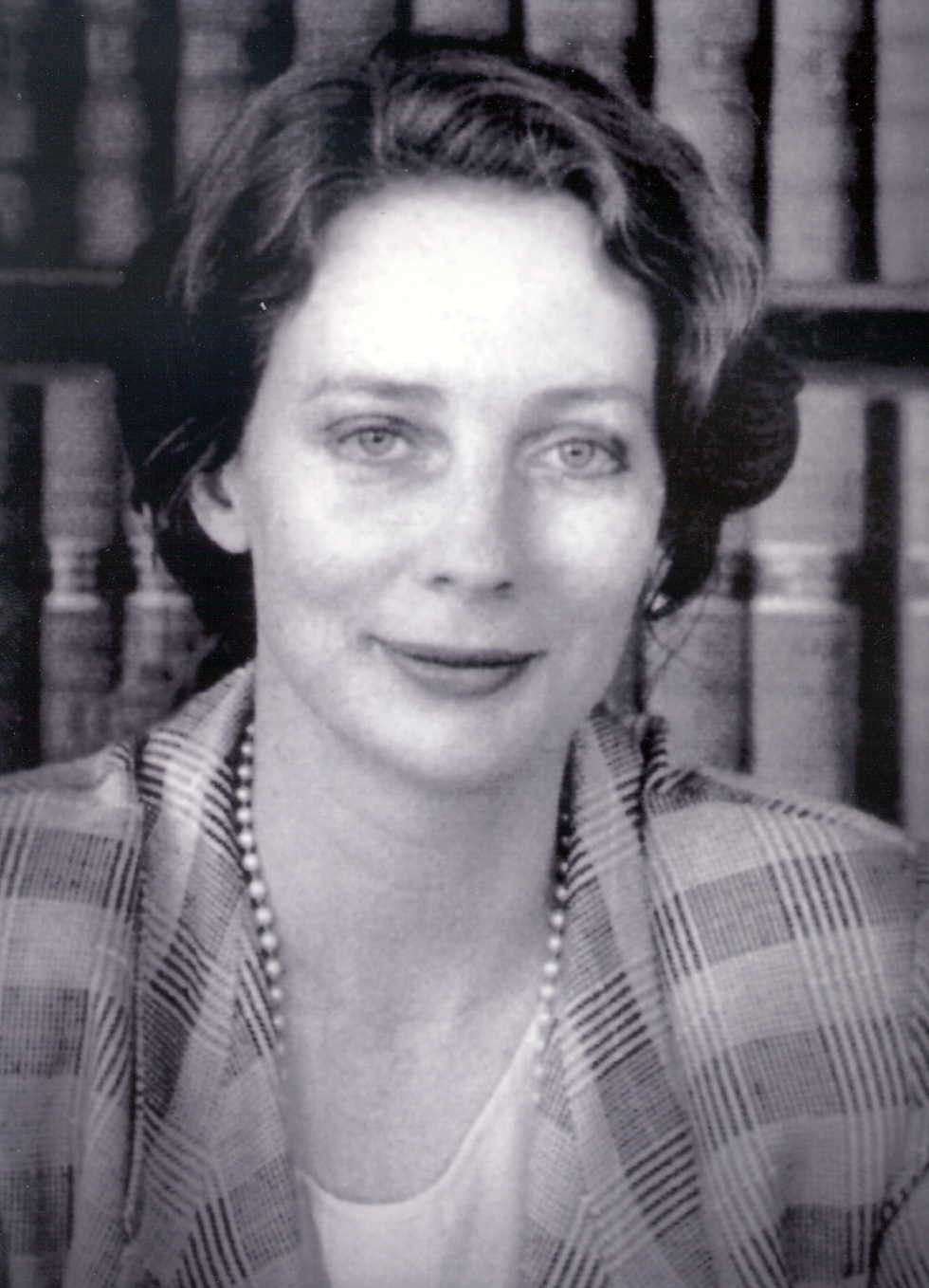 Black-and-white portrait of a smiling person in front of bookshelves, wearing a plaid jacket and necklace