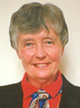 Portrait of a smiling person in a dark jacket, red shirt, and patterned tie on a plain background