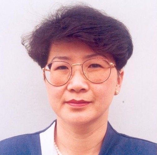 Portrait of a person with short dark hair and round glasses, wearing a blue jacket against a light background