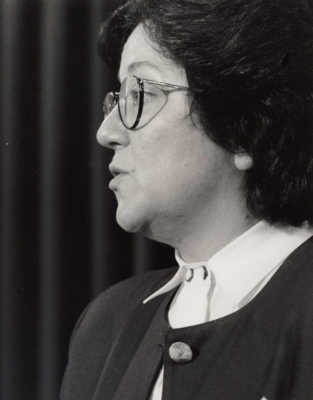 Side-profile portrait of a person with glasses and dark hair, wearing a dark jacket and white shirt