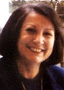 Smiling woman with shoulder-length dark hair outdoors, wearing a dark top and jacket.