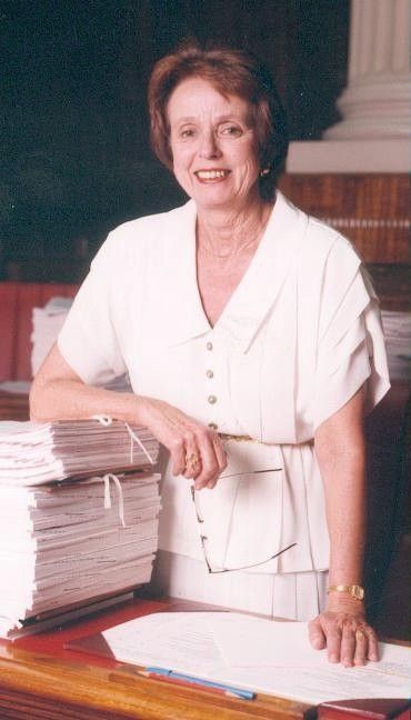 Woman in a white dress standing beside a tall stack of papers at a desk, smiling in an office.