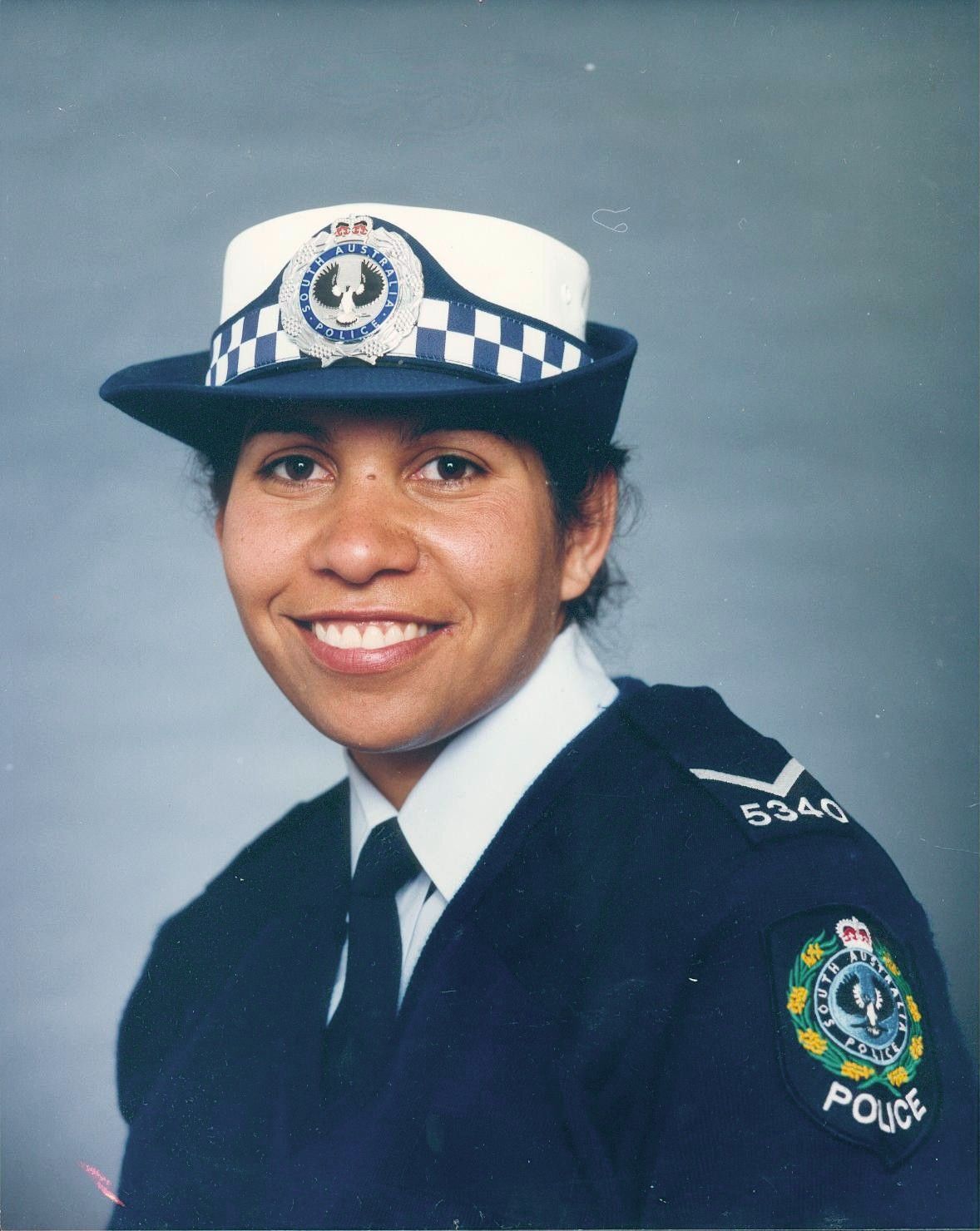 Police officer in dark uniform and white cap, smiling in a studio portrait.