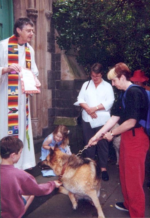 Clergy blessing a dog as people gather outdoors beside a stone building.