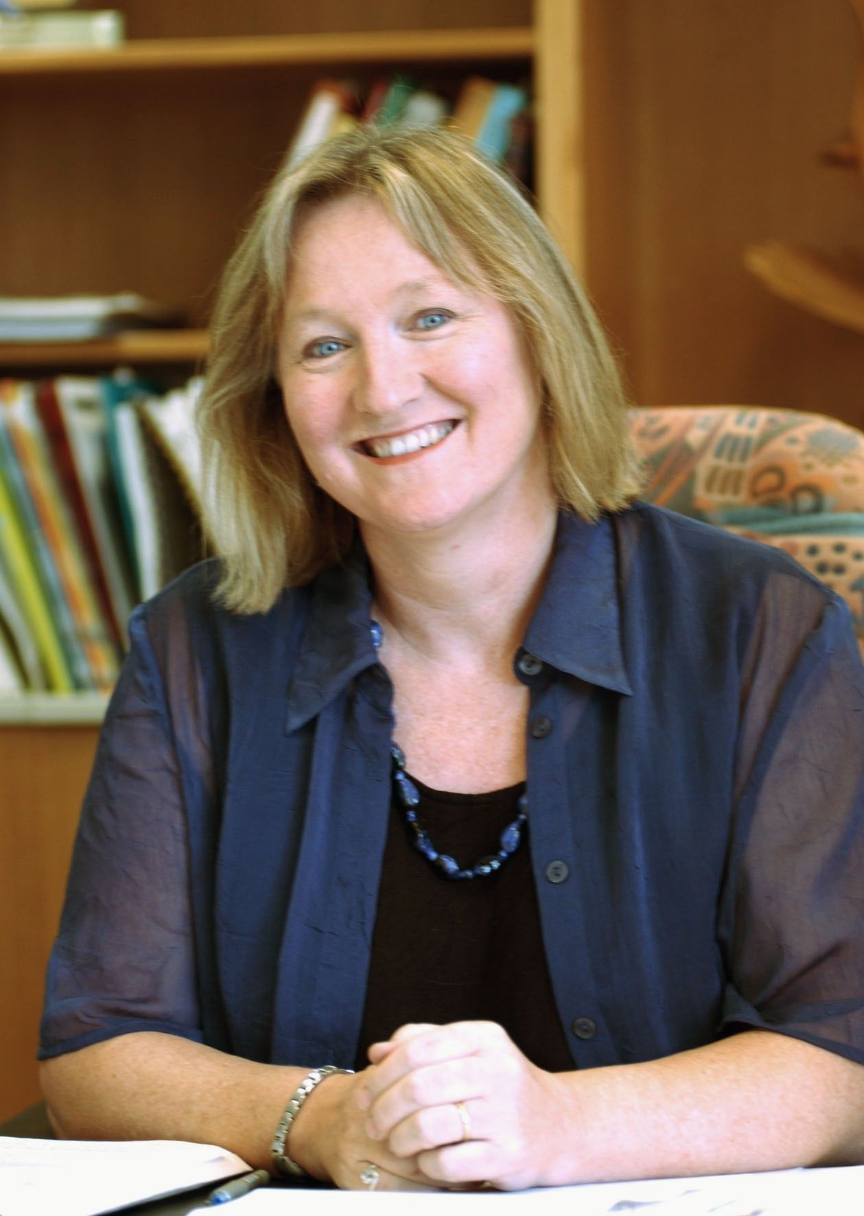 Smiling woman seated at a desk in an office, with bookshelves in the background.