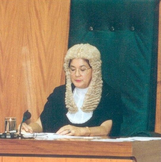 Person in a white judge’s wig sits at a desk in a courtroom, writing on papers.