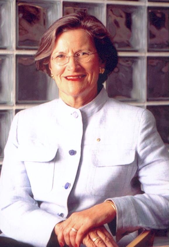 Portrait of a smiling woman in a white jacket, seated before a glass block wall.