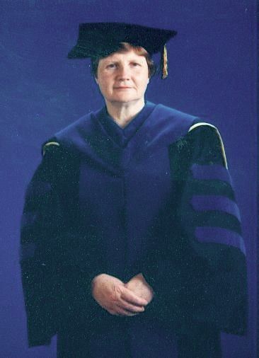 Person in graduation gown and cap, standing against a blue background with hands clasped.