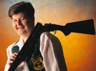 Smiling person holding a rifle over their shoulder against an orange background